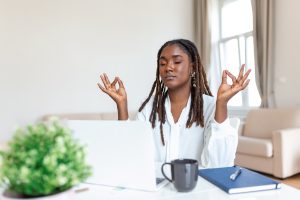 Calm African female executive meditating taking break at work for mental balance, mindful businesswoman feeling relief and no stress doing yoga at work ignoring avoiding stressful job for staff wellbeing purposes